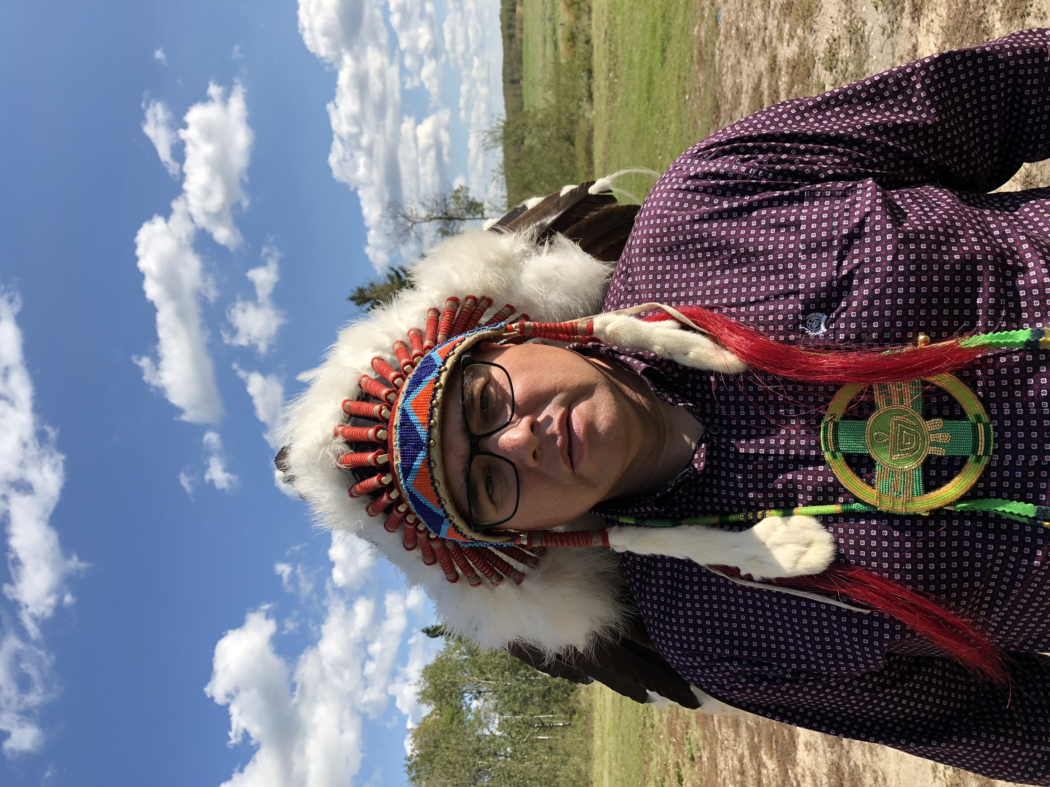 Delbert Wapass standing outdoors in a feathered headdress and beaded medallion
